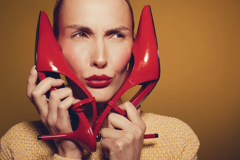 Woman holds red high heel shoes near face with thoughtful expression. Studio portrait on brown background. Concept of fashion, style, and footwear accessories for women.