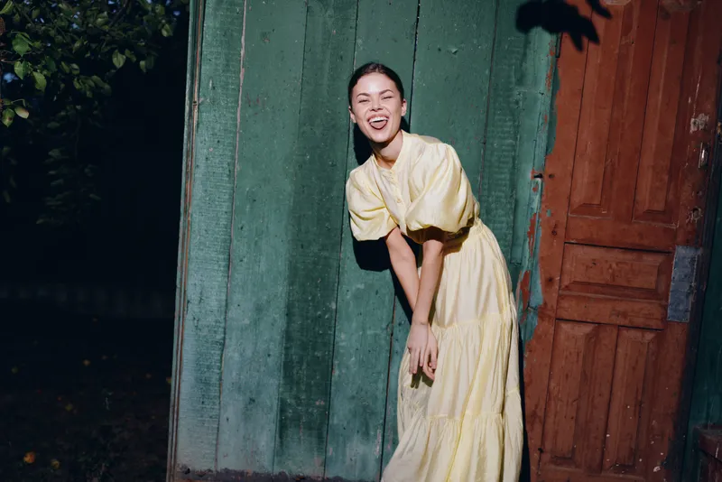 woman wearing a yellow dress posing by a doorway at night in an outdoor setting, with a candid smile and relaxed stance that conveys warmth and fashion flair
