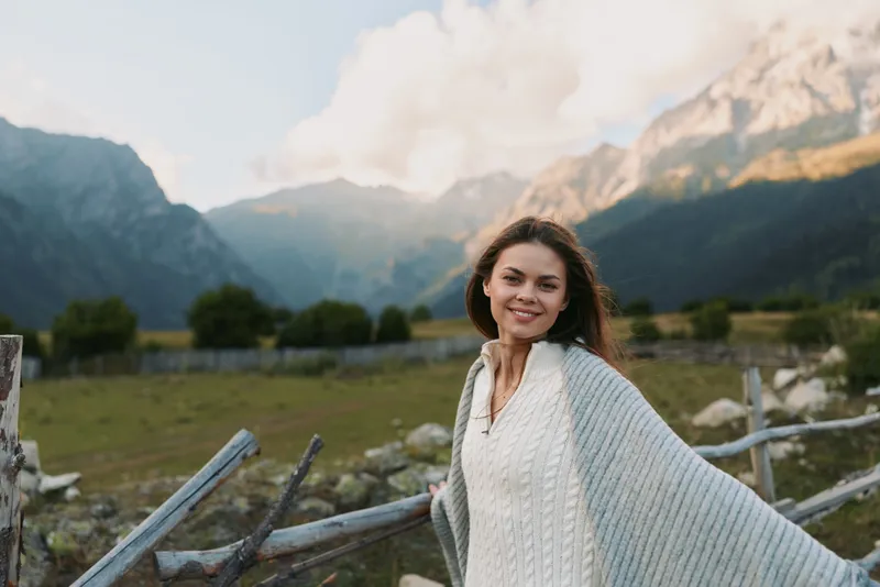 Smiling woman wrapped in a blanket standing by wooden fence in mountain landscape. Clear sky and natural light create peaceful outdoor lifestyle mood.
