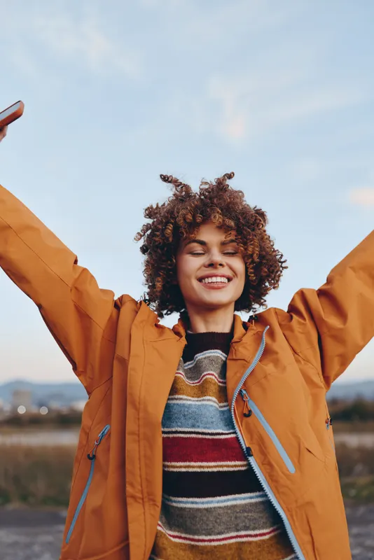 Cheerful woman outdoors, curly hair, smiling with arms raised, wearing orange jacket and striped sweater, warm sunset light, open field landscape, casual lifestyle, relaxed posture