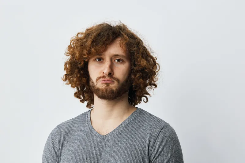 Young man with curly hair and a neutral expression, wearing a gray sweater in a minimalist setting, representing emotions and introspection