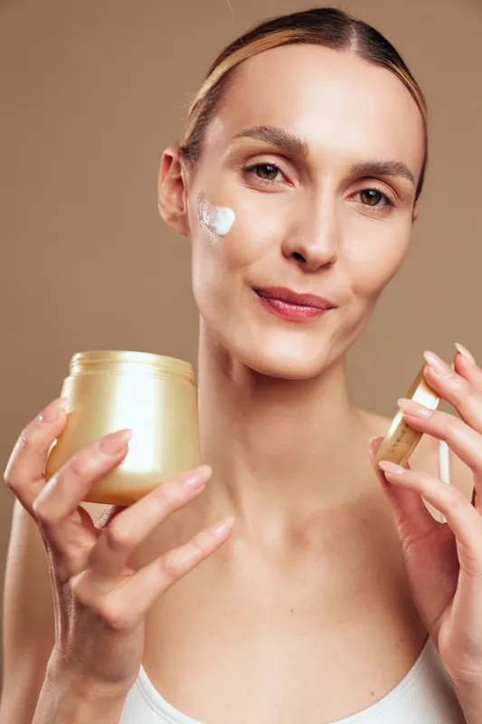 Young woman applies facial moisturizer, holding a cosmetic jar and touching her cheek, daily skincare beauty routine on warm neutral background, concept of self care, healthy skin glow