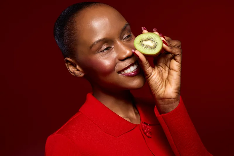 Smiling dark skinned African woman in red sweater holds half of kiwi fruit near face on red background. Concept for healthy lifestyle, natural beauty, fresh fruit, and nutrition.