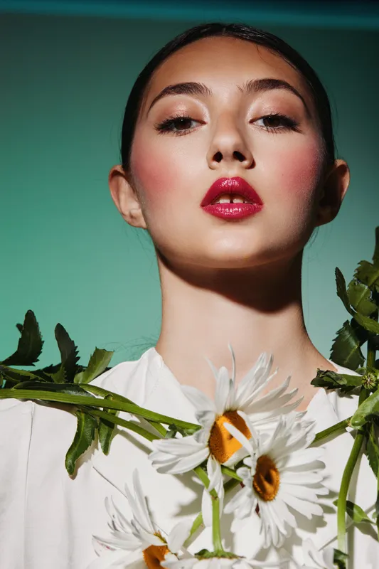 Close-up of confident young woman with glossy red lips and pink blush holding white daisies against turquoise background. Beauty fashion concept with minimal jewelry styling.