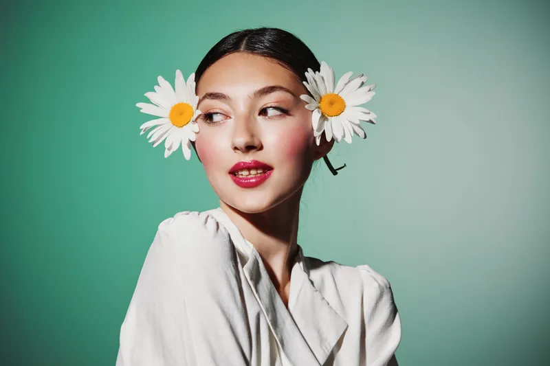 Young woman with red lipstick and floral earrings looks sideways with a smooth complexion. Artistic beauty and fashion concept on a soft turquoise background. Studio shot.