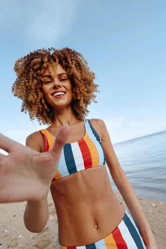 On a sunny beach by the ocean, a woman in a colorful swimsuit smiles, curly hair blowing as she waves at the camera, summer vacation mood, candid outdoor portrait.