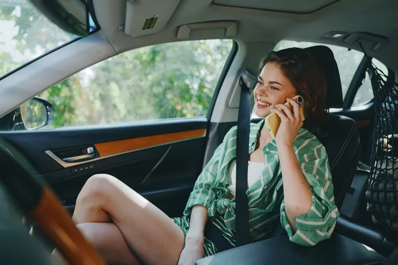 Young woman sitting in a car seat, smiling and talking on a smartphone, relaxed and stylish in casual attire.
