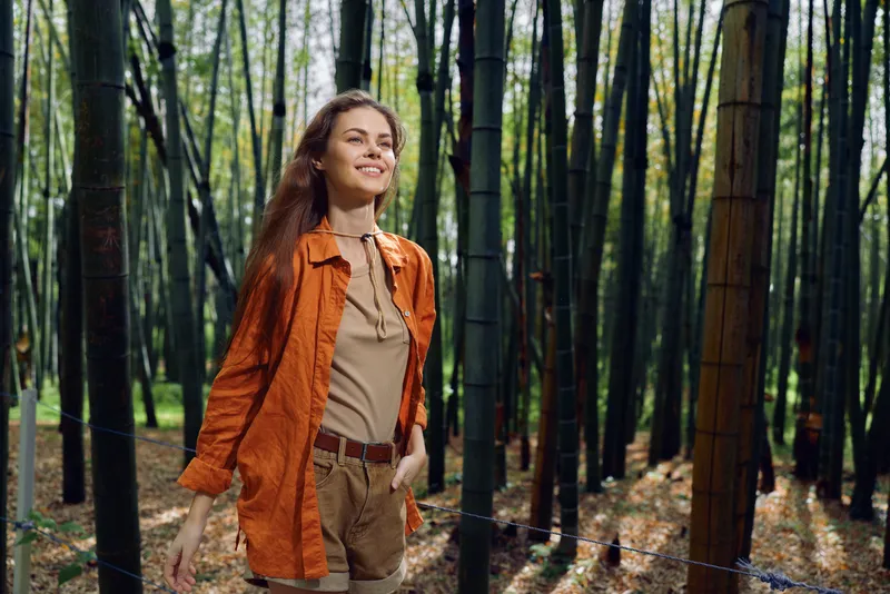 Woman walking in bamboo forest, smiling and enjoying nature outdoors in casual outfit. Young female traveler exploring tall bamboo grove with sunlight and peaceful green surroundings.
