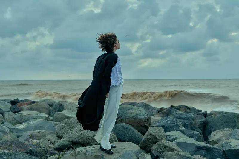 Woman standing on rocky shoreline with flowing coat, facing the sea under a moody sky, capturing wind, movement and dramatic ocean scenery