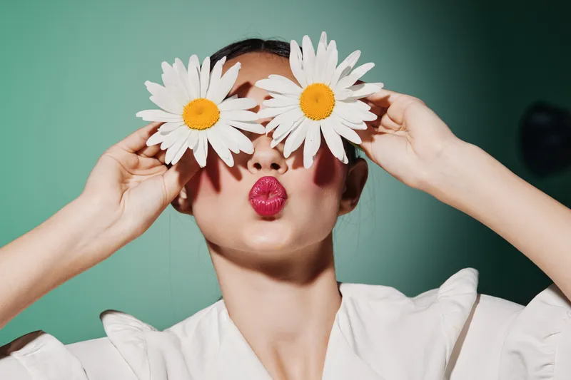 Creative woman holds large white daisies over eyes posing with bright red lips puckered. Studio portrait on green background. Concept of beauty fashion and playful mood.