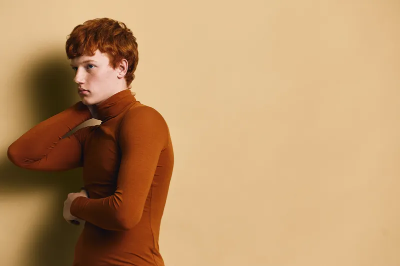 Young man with red hair and porcelain skin wearing brown turtleneck standing against beige wall with thoughtful expression looking away from camera in studio.