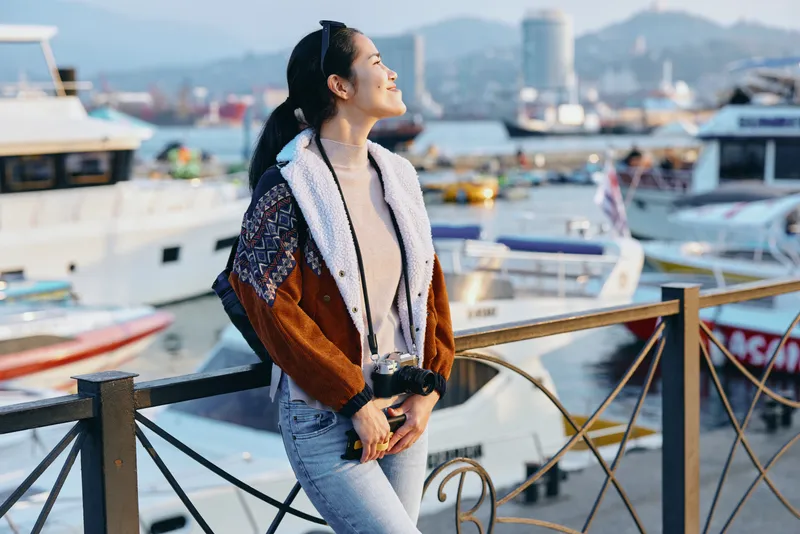 Woman in a stylish jacket stands at a harbor railing, looking toward boats bathed in afternoon light, capturing a moment of seaside leisure and travel curiosity by the marina, with gentle waves and
