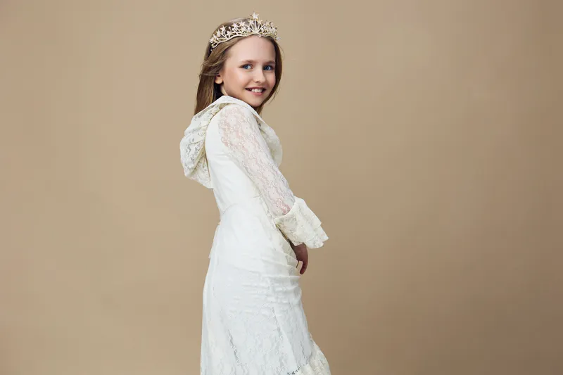 Charming young girl in a delicate white dress with a crown, smiling gracefully against a soft beige background, conveying joy and innocence in a serene setting