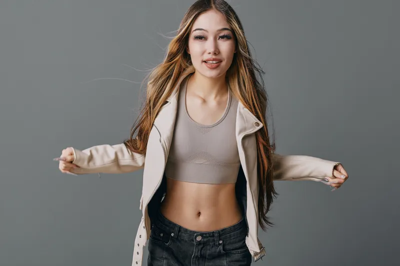 Young woman with long hair wearing casual crop top and jacket posing with arms outstretched on gray background. Fashion, beauty and confidence in studio portrait for lifestyle and style.