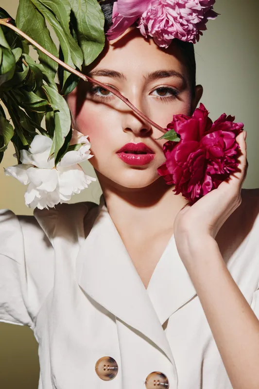 Beauty fashion portrait of young woman with bold red lips and floral arrangement wearing white blazer with shadows on face. Creative artistic studio beauty shot.