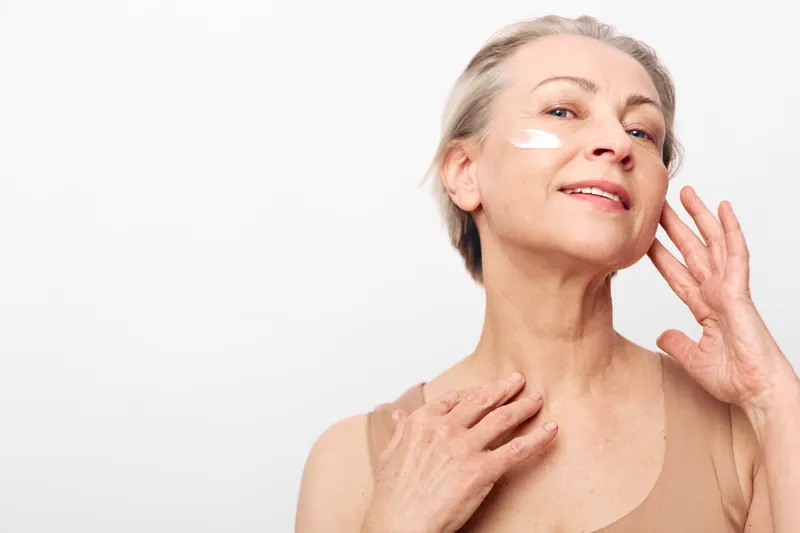 Mature woman with healthy skin applies white face cream on cheek, smiling softly. Skincare concept for aging care, beauty routine, hydration, and self-confidence in studio light.