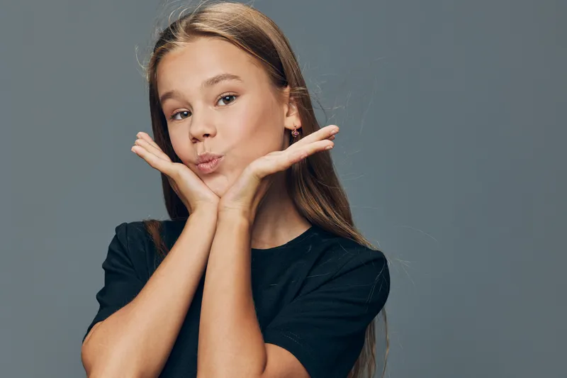 Girl with long hair making kiss face with hands near cheeks posing on gray background. Portrait of young female child showing playful and cute expression in studio.