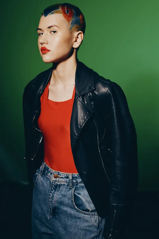 Woman with colorful short hair wearing a black leather jacket and red top, standing confidently against a green background, showcasing bold style and attitude.
