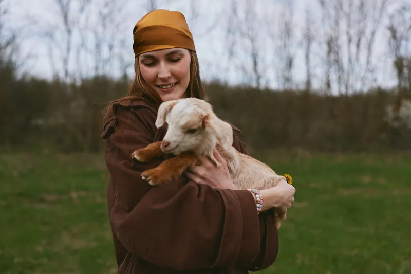 Young woman wearing a brown robe and orange scarf smiles while cradling a small goat kid in her arms in a green field, depicting gentle companionship and a warm rural lifestyle.