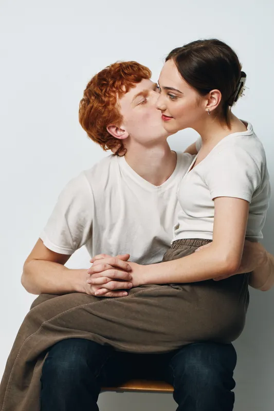 Couple, love, kiss and affection with young man and woman sitting closely with hands held on chair for romantic connection and intimacy in studio with white background and casual clothes.