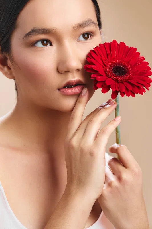 Asian woman gazes softly while holding a red flower close to her face, creating an intimate studio portrait that emphasizes beauty, calm, and elegant, gentle mood in a clean setting.