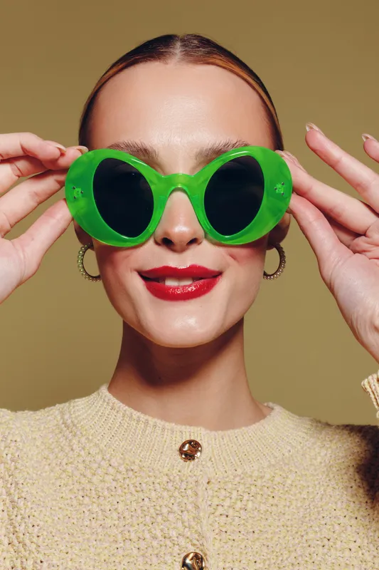 Young woman with slicked hair wears oversized green sunglasses and red lipstick, smiling confidently against beige background. Concept of fashion, style, and summer accessories.