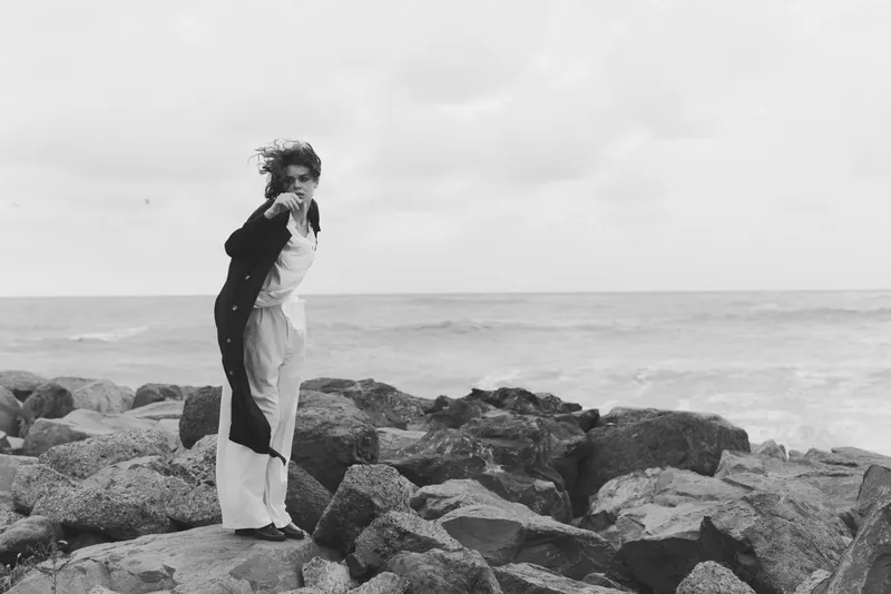Woman stands on rugged rocks by the sea, wearing a light dress and dark coat, wind in her hair, monochrome coastal scene conveying resilience and solitude in seaside moment for stock