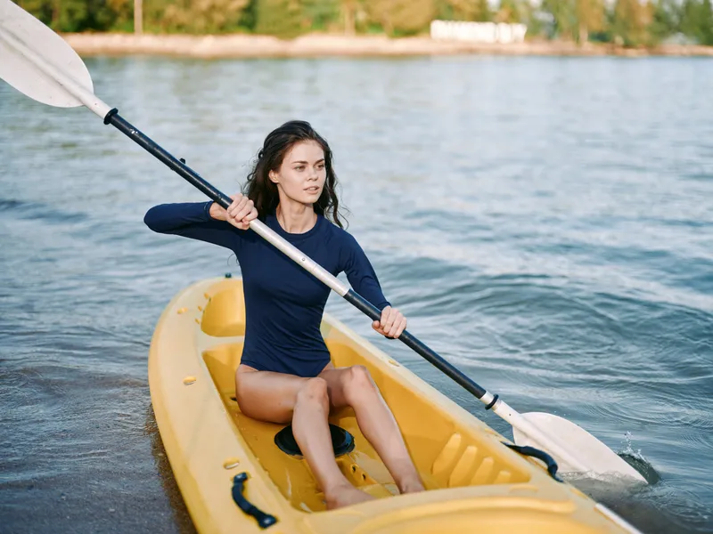 Focused young woman paddles a bright yellow kayak along calm water, gripping a double blade oar with steady rhythm and confident posture as the scene unfolds.