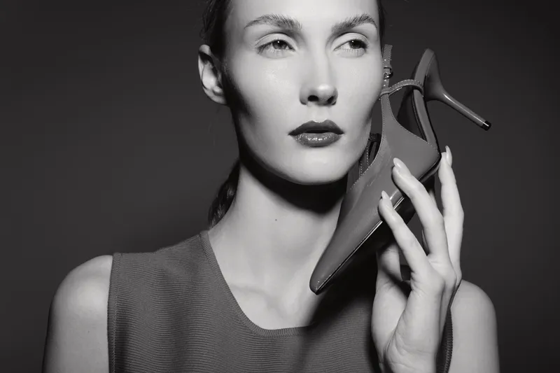 White woman with fair skin holds high heel shoe near face in black and white studio portrait. Concept of fashion, style, elegance, and beauty with artistic lighting.