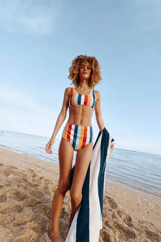 Woman beach ocean swimsuit striped towel curly hair summer sunlight relaxed walk along sandy shore, gentle waves, clear blue sky, warm natural colors, carefree vacation mood