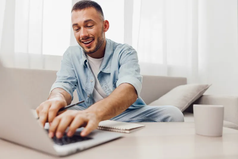 Smiling man working from home, dressed in casual attire, focused on his laptop. Bright interior with soft natural light creating a cozy atmosphere.