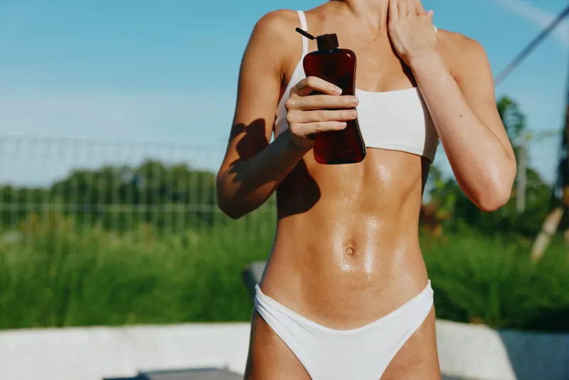 Tanned woman in white bikini applying sunscreen lotion on body outdoors. Close-up of smooth skin with sun protection concept, summer lifestyle, and healthy skin care under natural sunlight.