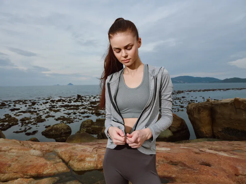 Young athletic woman in activewear striking a thoughtful pose on rocky shore, embodying fitness and determination amidst serene coastal backdrop