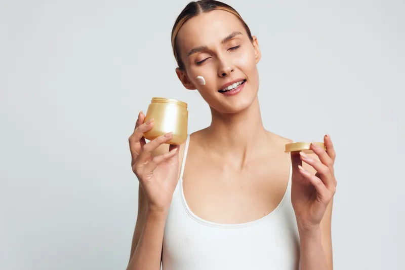 Young woman with blonde hair applies moisturizer from a gold jar, smiling softly, wearing a white tank top, standing against a clean light gray background, skincare routine in progress