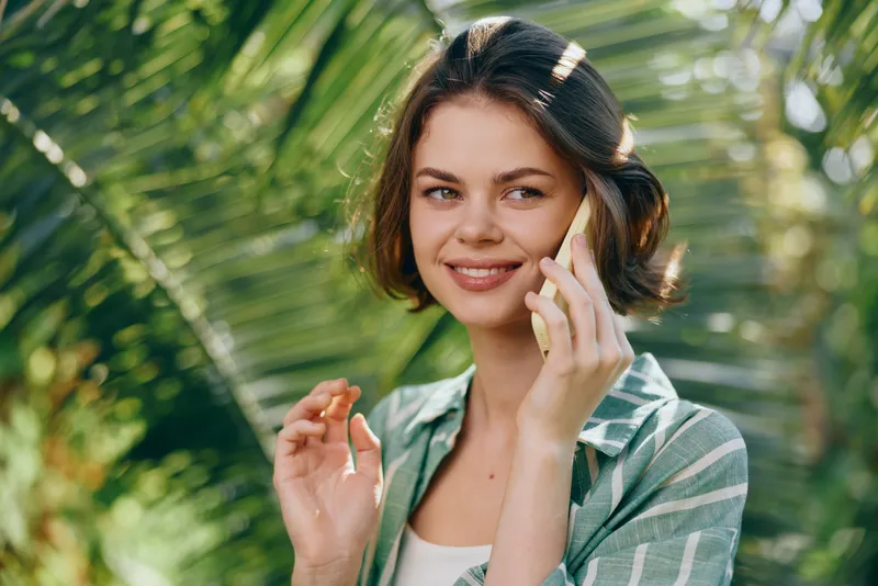 A smiling woman talks on a phone outdoors amid lush greenery, wearing casual clothing with natural light highlighting her friendly expression, conveying everyday communication in a sunny setting.
