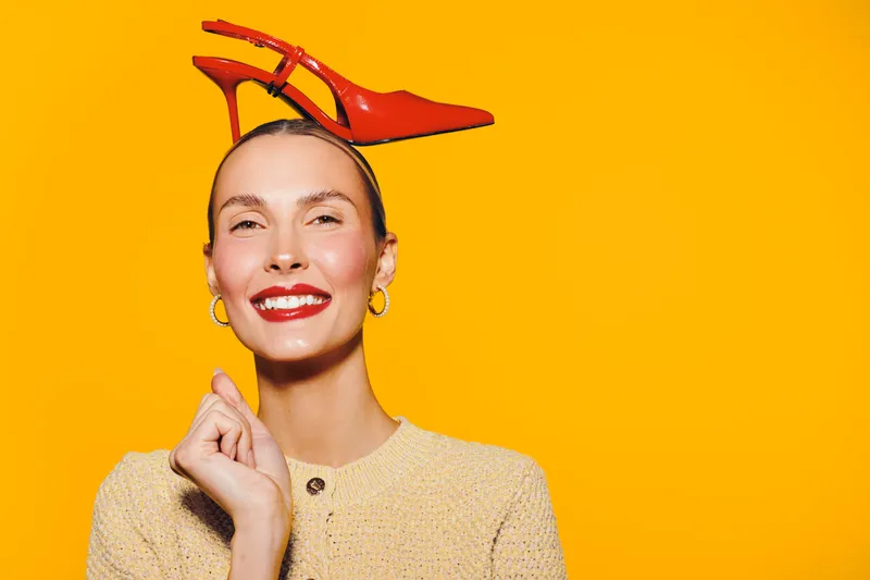 Smiling woman with red lipstick holds red high heel shoe on head against bright yellow background. Concept of fashion, style, beauty, and playful mood in studio portrait.