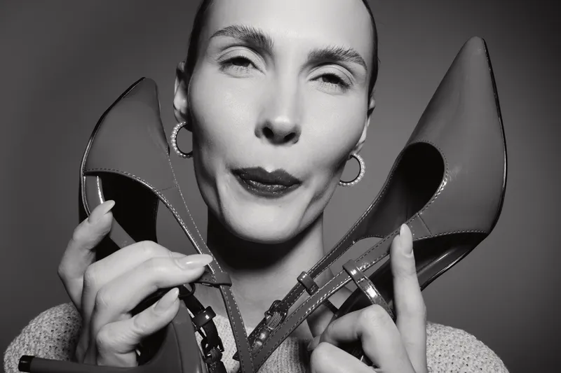 Close-up portrait of a light skinned Caucasian woman holding high heels near her face, wearing hoop earrings and a cozy sweater in a monochrome studio. Concept of fashion and style.