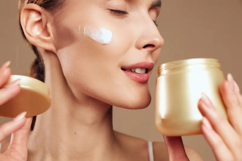 Smiling woman applying facial cream from a jar. Close-up on a warm beige background. Skincare, wellness, and self-care concept. Soft studio lighting creates a gentle, uplifting mood.