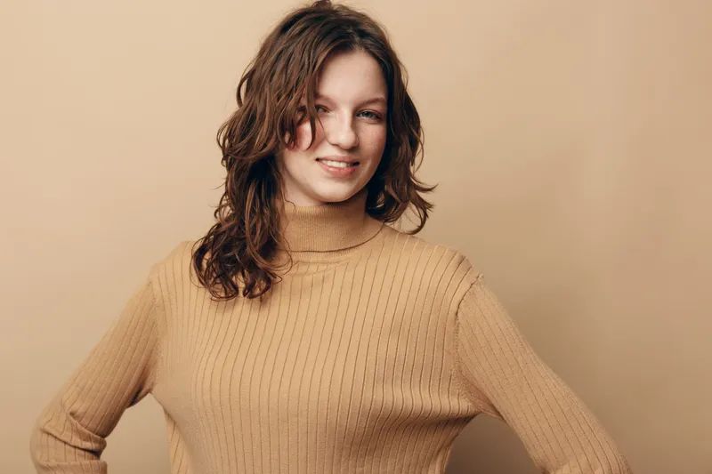 Smiling young woman with curly brown hair wearing a beige knitted sweater stands confidently against a plain beige background. Concept of casual style and natural beauty.