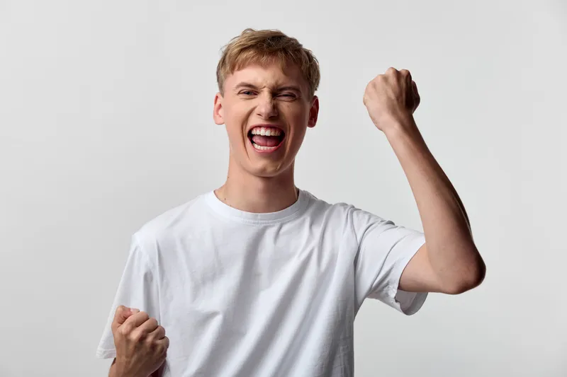 Young man in a white t shirt celebrating with raised fists and a wide smile, conveying excitement, victory and high energy in a simple studio setting