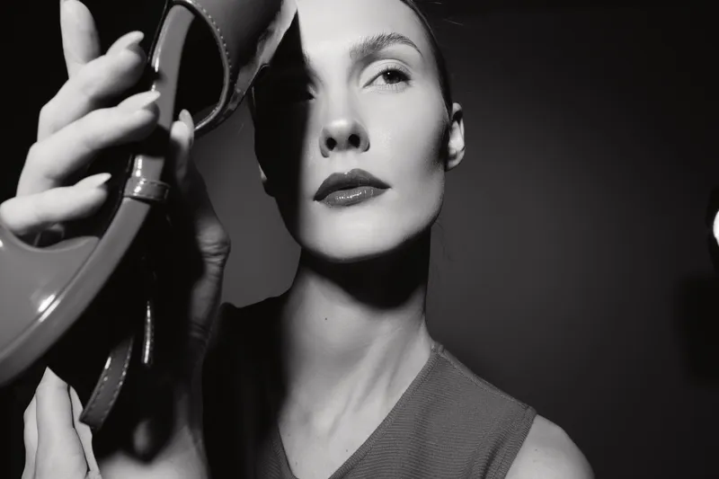 Elegant white woman with light skin holds a shoe near her face in black and white studio portrait. Concept of fashion, style, and minimalism.