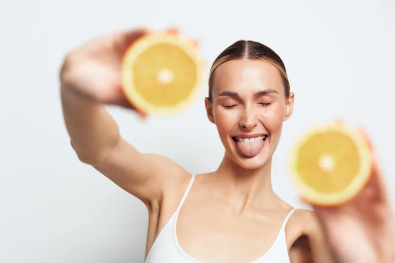 A playful young woman with hair tied back holds two halves of a fresh orange close to the camera while sticking out her tongue with eyes closed on a white background.
