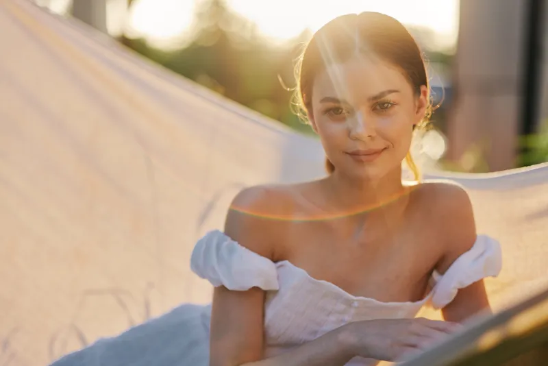 Young woman in a white dress relaxing on a hammock during sunset, evoking serenity and joy in a warm, natural outdoor setting