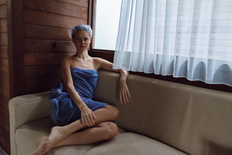young woman wrapped in towel relaxing on beige sofa by window in cozy wooden room, natural light, calm atmosphere, minimalistic interior, casual pose