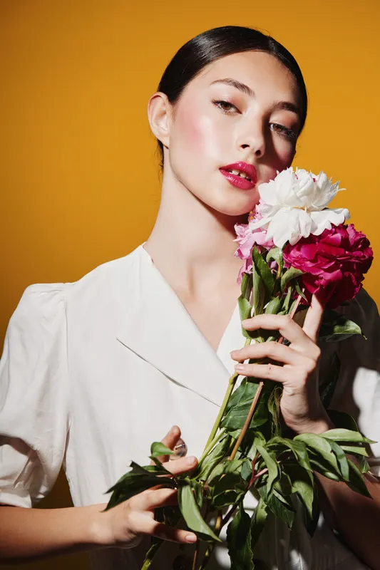 Elegant woman with glossy skin and bright lips holds colorful flowers against a vibrant orange background. Studio portrait with high-fashion beauty and creative concept.