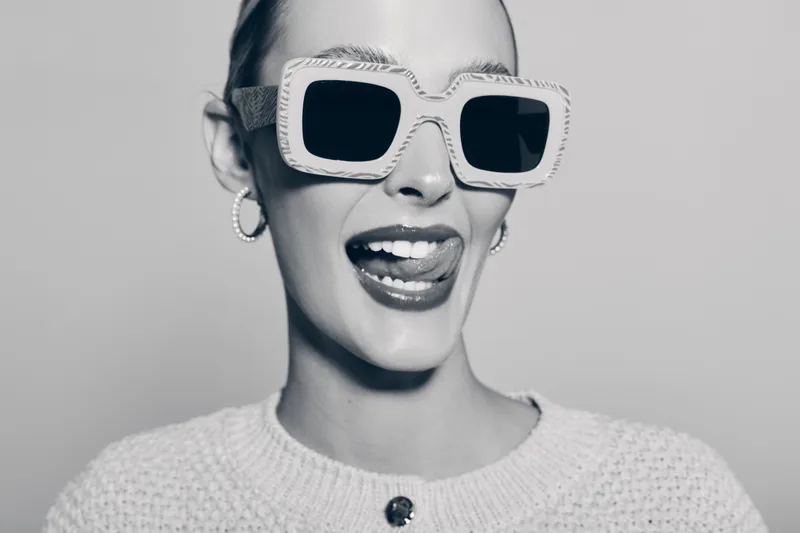 Young white woman with fair skin wearing oversized sunglasses and hoop earrings smiles playfully with her tongue out. Black and white studio portrait with casual sweater.