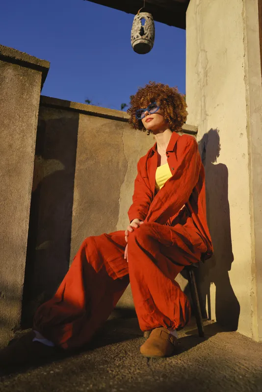 Cheerful woman outdoors wearing a red jumpsuit, seated on a sunlit terrace, relaxed pose, soft breeze, natural tones, urban backdrop, stylish lifestyle moment