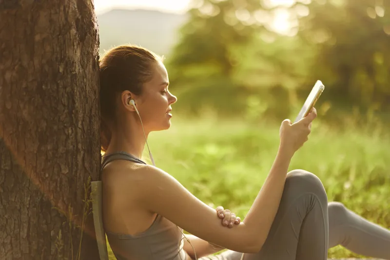 Young athletic woman enjoying music on her smartphone while sitting against a tree in a sunny park, illustrating relaxation and leisure activities