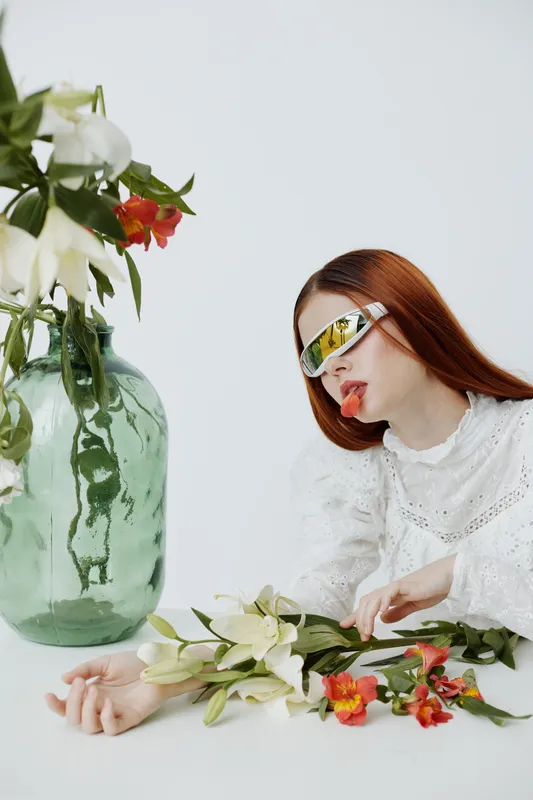 Stylish female model with long red hair wearing futuristic sunglasses, lying next to a vase of flowers in a minimalistic setting