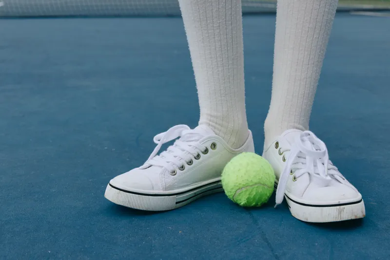 Young athlete s feet in white sneakers on a tennis court, with a vibrant green tennis ball, showcasing energy and sportiness in a bright and active environment